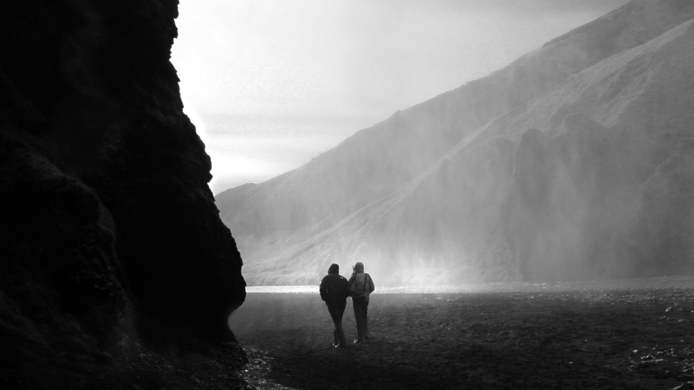 Due viandanti camminano insieme in una valle, tra una parete di roccia scura e la luce nella nebbia